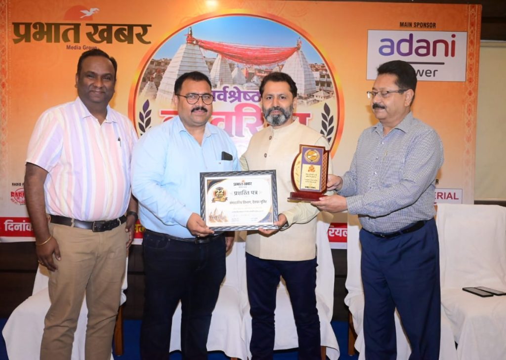 prabhat khabar deoghar | No. 1 Indian Media News Portal Four men stand on a stage during an awards ceremony; one hands a framed certificate and another holds a wooden trophy, with a bright event banner in the background.