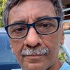 Close-up portrait of a middle-aged man with blue-framed glasses, gray hair and mustache, wearing a light blue shirt outdoors.