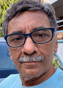 Close-up portrait of a middle-aged man with blue-framed glasses, gray hair and mustache, wearing a light blue shirt outdoors.