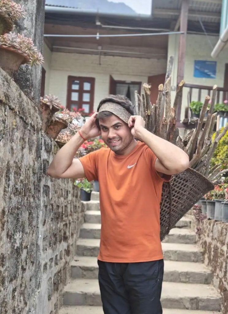 prakash badal | No. 1 Indian Media News Portal Smiling man in an orange shirt adjusts a headband while carrying a woven basket of branches along a stone wall.