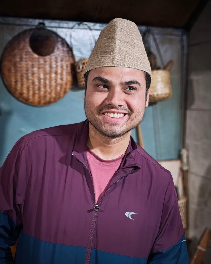 Smiling man in a maroon jacket with a beige woven cone hat, standing in a room with wicker baskets in the background.