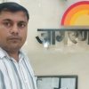 A man posing for a selfie beside a wall sign with a rainbow semicircle logo and metallic letters.