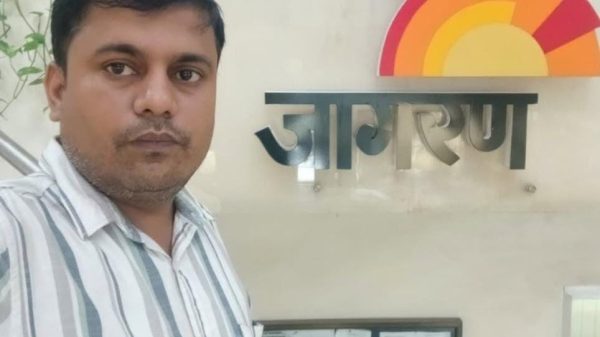 A man posing for a selfie beside a wall sign with a rainbow semicircle logo and metallic letters.
