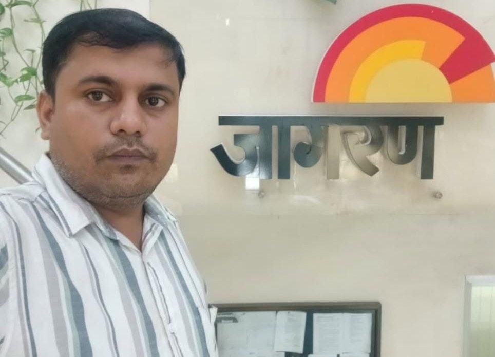 A man posing for a selfie beside a wall sign with a rainbow semicircle logo and metallic letters.