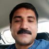 Close-up of a man with a dark mustache in a car, looking at the camera with a neutral expression.