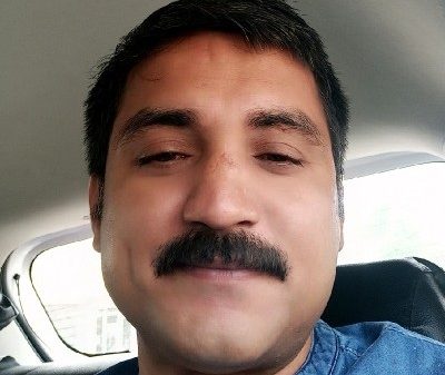 Close-up of a man with a dark mustache in a car, looking at the camera with a neutral expression.