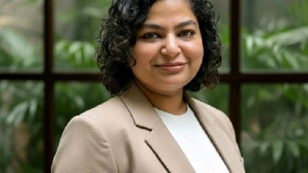 Portrait of a woman with short curly hair, wearing a beige blazer and white top, smiling in front of a glass window with greenery behind her.