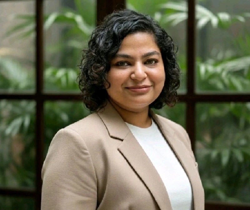 Portrait of a woman with short curly hair, wearing a beige blazer and white top, smiling in front of a glass window with greenery behind her.