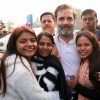 Smiling group outdoors: a bearded man in a white polo shirt at center, embraced by four women, with a crowd and a car in the background.