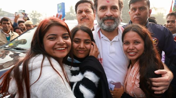 Smiling group outdoors: a bearded man in a white polo shirt at center, embraced by four women, with a crowd and a car in the background.