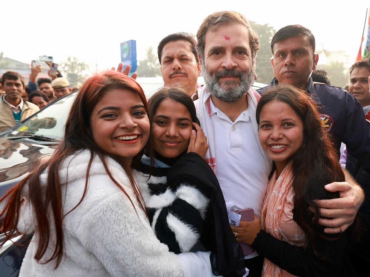 Smiling group outdoors: a bearded man in a white polo shirt at center, embraced by four women, with a crowd and a car in the background.
