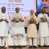 Eight men stand on a stage during a book launch, each holding a copy of the same book; a large poster with a portrait and Hindi text serves as the backdrop.