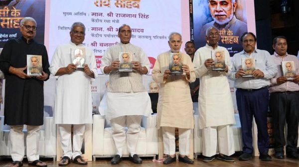 Eight men stand on a stage during a book launch, each holding a copy of the same book; a large poster with a portrait and Hindi text serves as the backdrop.