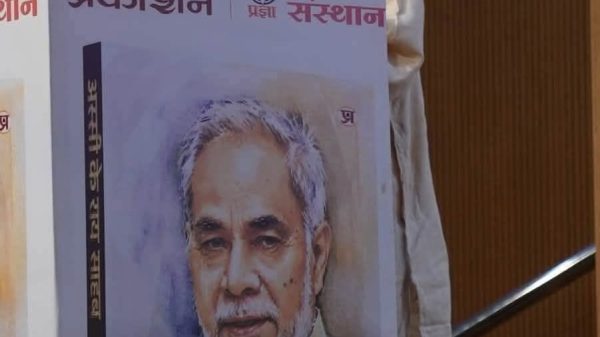 An elderly man speaks at a podium next to a large poster showing his portrait on a book cover.