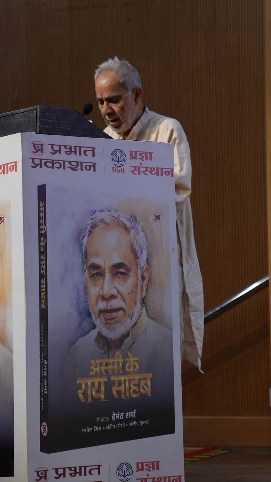 An elderly man speaks at a podium next to a large poster showing his portrait on a book cover.