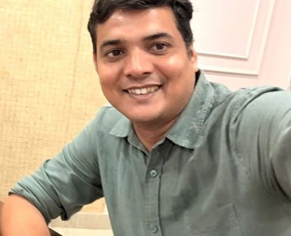 Smiling man with short dark hair wearing a teal-green shirt, taking a selfie indoors at a table.