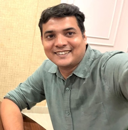 Smiling man with short dark hair wearing a teal-green shirt, taking a selfie indoors at a table.
