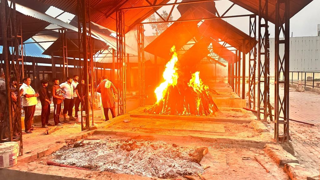 Line of people standing along a metal-framed shelter watching a large bonfire burning in a dirt yard under a covered area.”