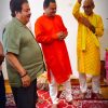 Three men in traditional Indian outfits at a ceremony, standing barefoot on patterned carpets under hanging garlands.