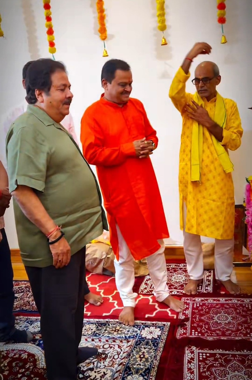 Three men in traditional Indian outfits at a ceremony, standing barefoot on patterned carpets under hanging garlands.