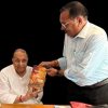 Two Indian men in white shirts, one seated and one standing, exchange a yellow-orange snack bag against a black background.