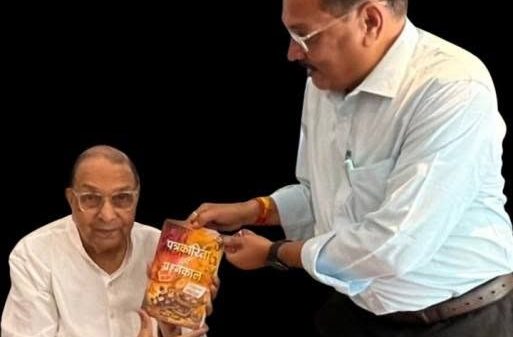Two Indian men in white shirts, one seated and one standing, exchange a yellow-orange snack bag against a black background.