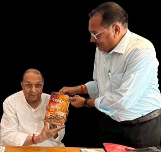 Two Indian men in white shirts, one seated and one standing, exchange a yellow-orange snack bag against a black background.