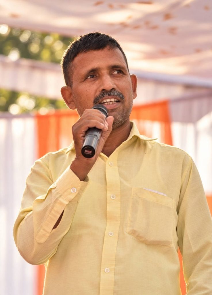 Man in a pale yellow shirt speaking into a handheld microphone at an outdoor event.