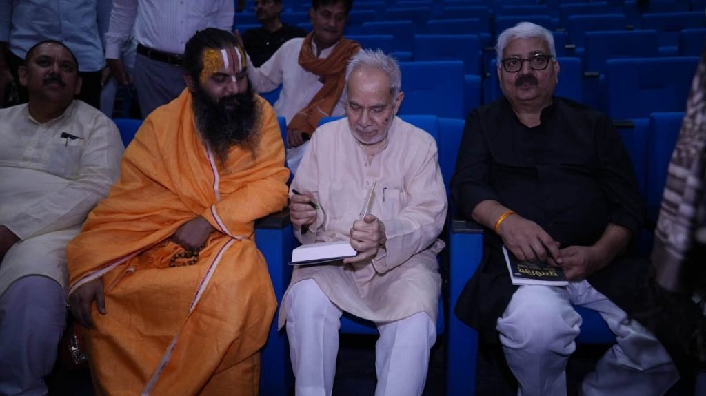 rb rai | No. 1 Indian Media News Portal Three men seated in blue theater chairs; a bearded swami in orange robes sits with the center man writing in a notebook; another man in black sits to the right with a book in his lap.