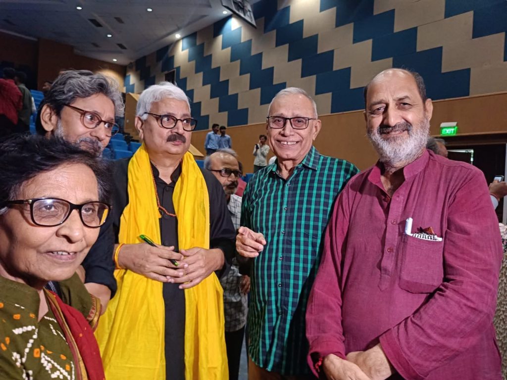 rb rai_2 | No. 1 Indian Media News Portal Five older men pose for a selfie in a conference hall with blue geometric-patterned walls; one wears a bright yellow scarf and holds a green pen.