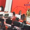 Newsroom scene with police officers escorting a person through a row of computer desks and monitors in a red-themed studio backdrop, TV logo in corner.