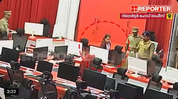 Newsroom scene with police officers escorting a person through a row of computer desks and monitors in a red-themed studio backdrop, TV logo in corner.