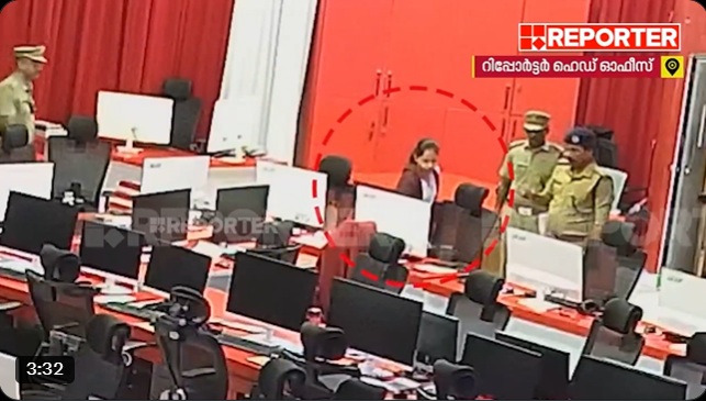 Newsroom scene with police officers escorting a person through a row of computer desks and monitors in a red-themed studio backdrop, TV logo in corner.