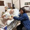 Elderly man in a hospital bed smiling as a younger person in a blue jacket reads him a colorful children's book from close up.