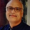 Portrait of a smiling bald man wearing glasses and a dark collared shirt, indoors.