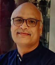Portrait of a smiling bald man wearing glasses and a dark collared shirt, indoors.
