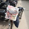 Stack of Hindi newspapers tied to a bicycle rack with red string on a street lined with parked motorcycles.