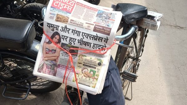 Stack of Hindi newspapers tied to a bicycle rack with red string on a street lined with parked motorcycles.
