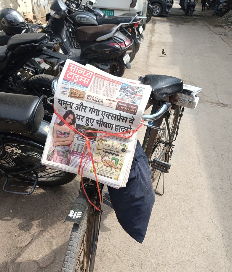 Stack of Hindi newspapers tied to a bicycle rack with red string on a street lined with parked motorcycles.
