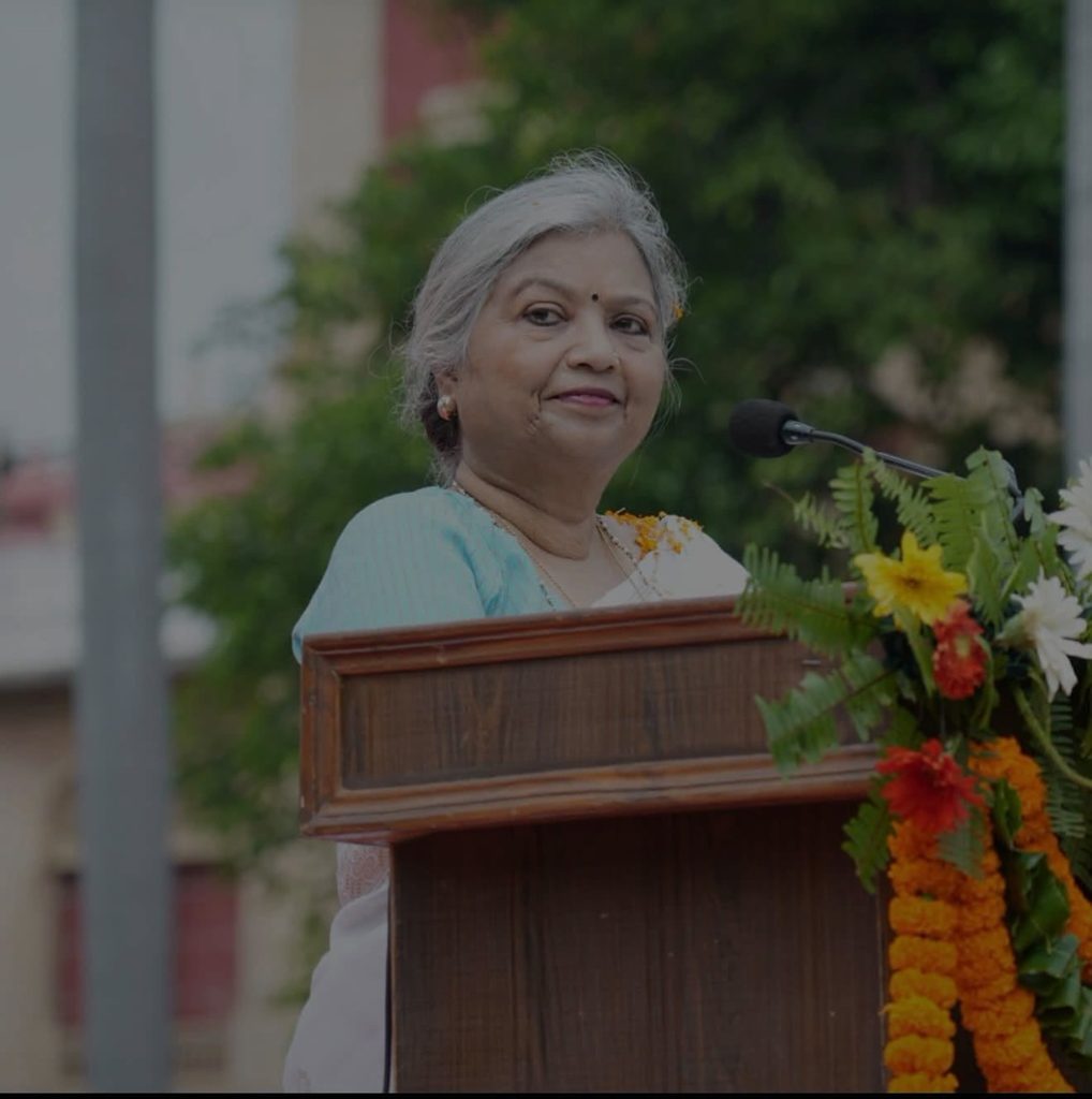 Screenshot | No. 1 Indian Media News Portal Older woman standing at a wooden podium with a microphone, delivering a speech; floral arrangement at the front. (Outdoor event)