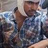 Man in a wheelchair with a head bandage and a bloodstained plaid shirt, in a hospital or clinic setting with medical staff in the background.