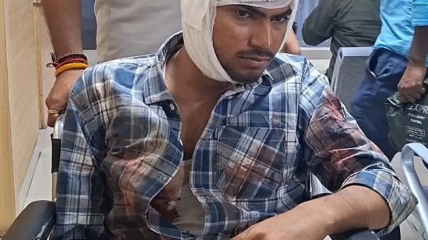 Man in a wheelchair with a head bandage and a bloodstained plaid shirt, in a hospital or clinic setting with medical staff in the background.