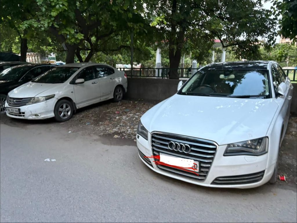 Two white cars parked side by side in a shaded lot beneath trees; the car in front is an Audi with a chrome grille.