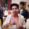 Center-frame man in a light-colored shirt unbuttoned at the chest, speaking to the camera indoors, with two people nearby in the background.