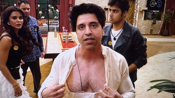 Center-frame man in a light-colored shirt unbuttoned at the chest, speaking to the camera indoors, with two people nearby in the background.