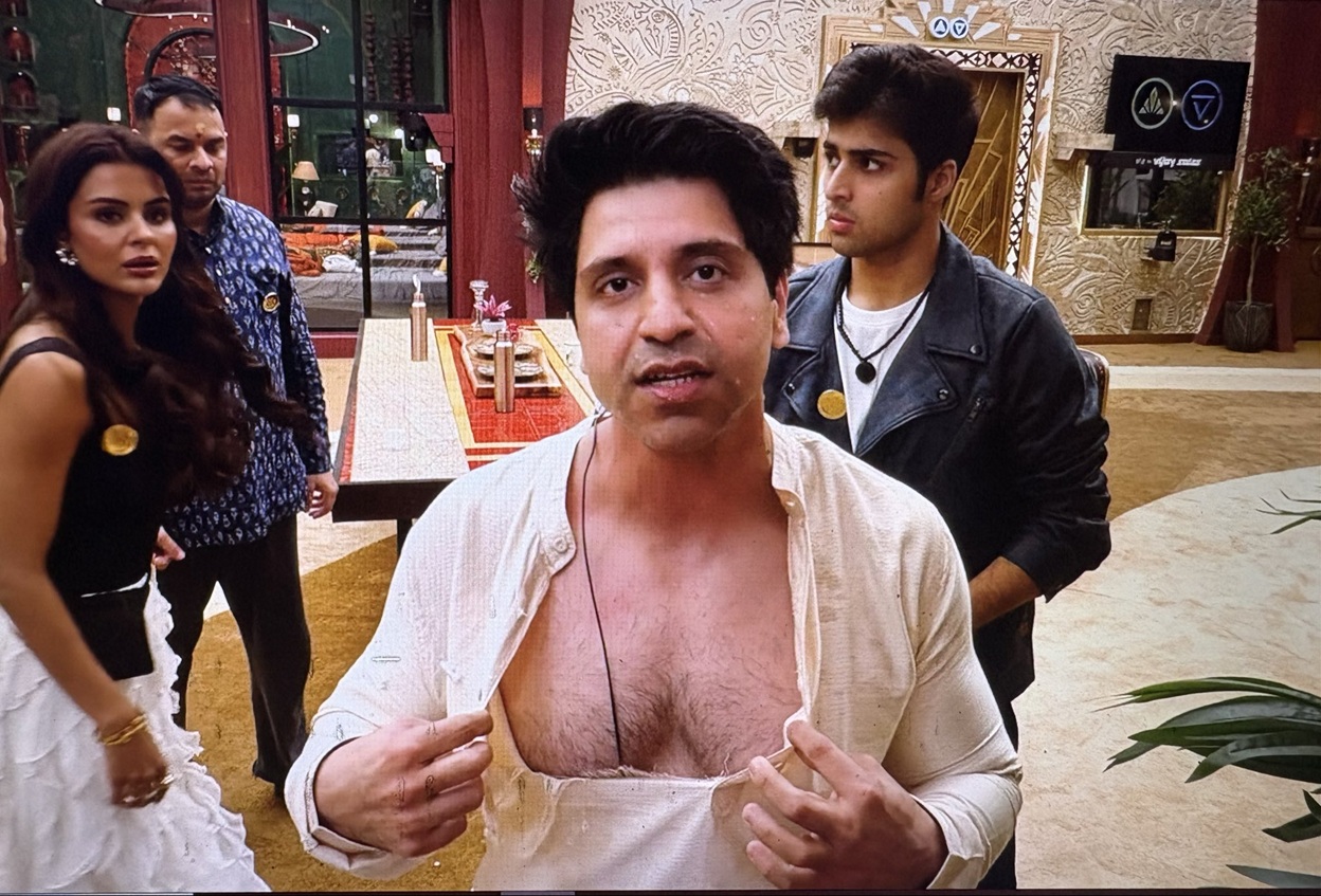 Center-frame man in a light-colored shirt unbuttoned at the chest, speaking to the camera indoors, with two people nearby in the background.