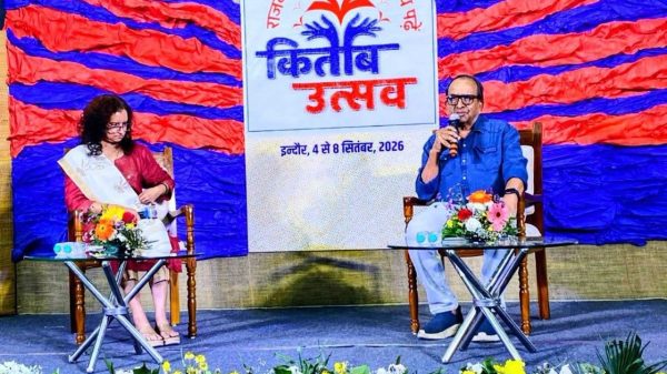 Man speaking into a microphone on stage, seated beside a woman in a pink outfit; colorful backdrop and festival banner with flowers behind them.