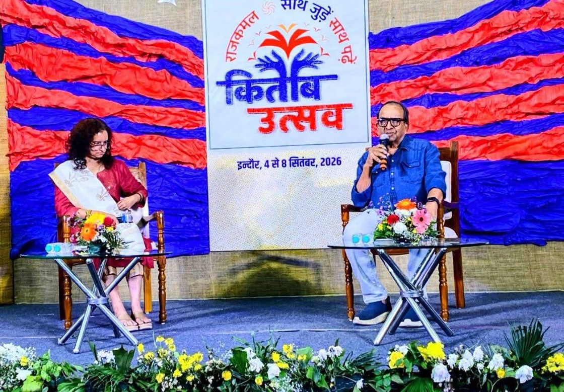 Man speaking into a microphone on stage, seated beside a woman in a pink outfit; colorful backdrop and festival banner with flowers behind them.