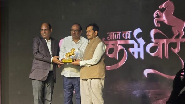 Three men on a stage exchanging a golden horse trophy at an award ceremony, with a dark backdrop featuring a pink horse design.