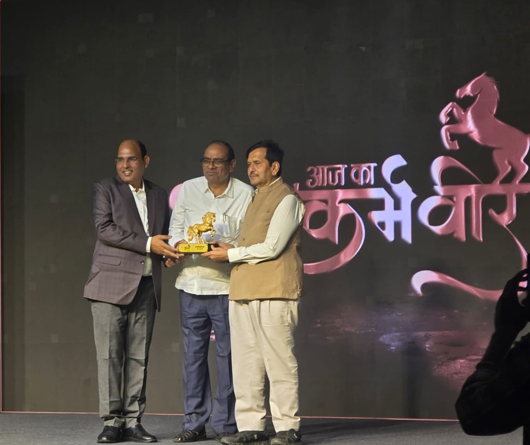 Three men on a stage exchanging a golden horse trophy at an award ceremony, with a dark backdrop featuring a pink horse design.